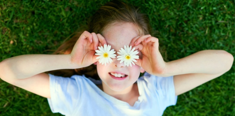 A top view of a young girl lying in the grass, holding daisies over her eyes