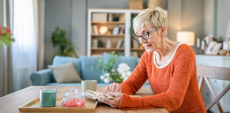 A mature woman sat at a table wearing glasses and reading a book.