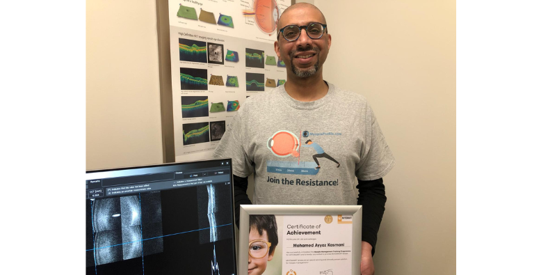 Mohamed Kasmani in Feltham Eyecare Centre wearing a Myopia Profile t-shirt, holding a Certificate of Achievement for HOYA MiYOSMART lenses, alongside a computer screen showing images of the eye.]