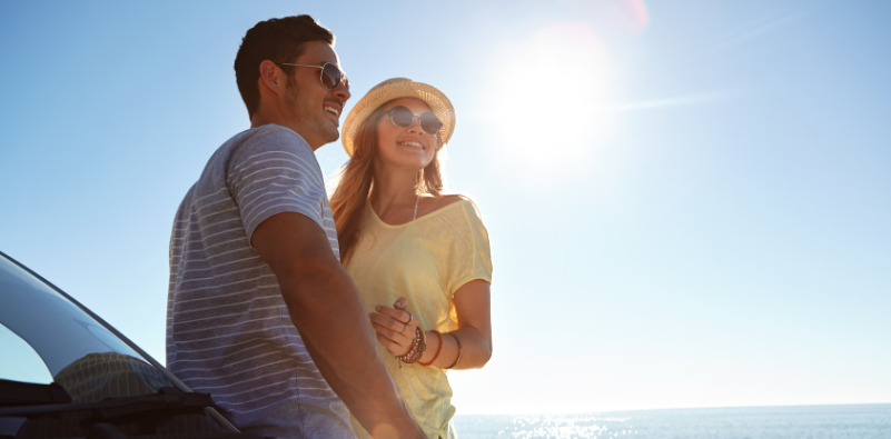A couple stood in front of their car wearing sunglasses beside a sunny beach